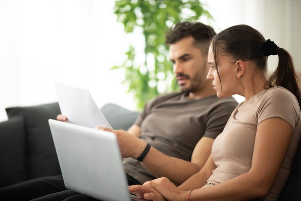 A couple sitting on a couch, looking at a laptop, submitting a cash offer request for their home.
